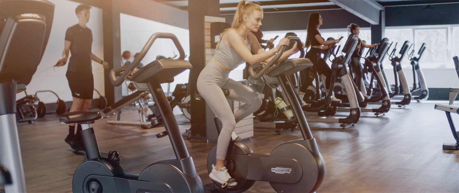 Lifestyle Szene im First X Fitnessstudio Dudweiler, Protagonistin auf dem Cardio Bike im Premium Studio Ambiente mit weiteren trainierenden Menschen im Hintergrund